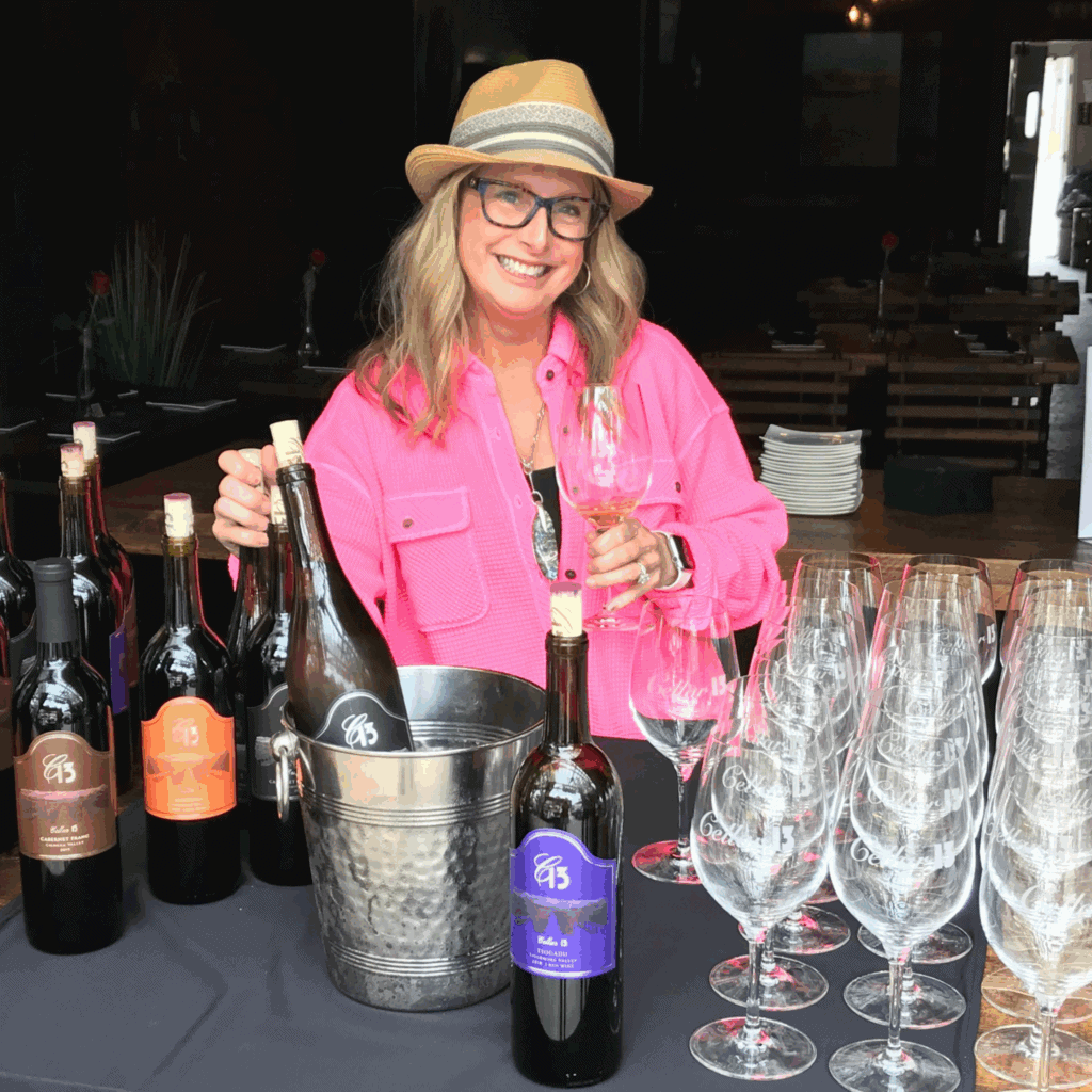 A picture of Gail Henderson of Cellar 13 Winery. She is pouring wine at an event and is wearing a hat and a bright pink shirt. 
