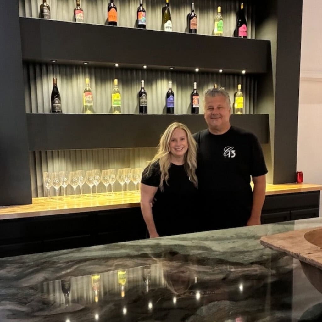 picture of Darin and Gail in front of shelving unit lit up with single bottles of Cellar 13 Winery.