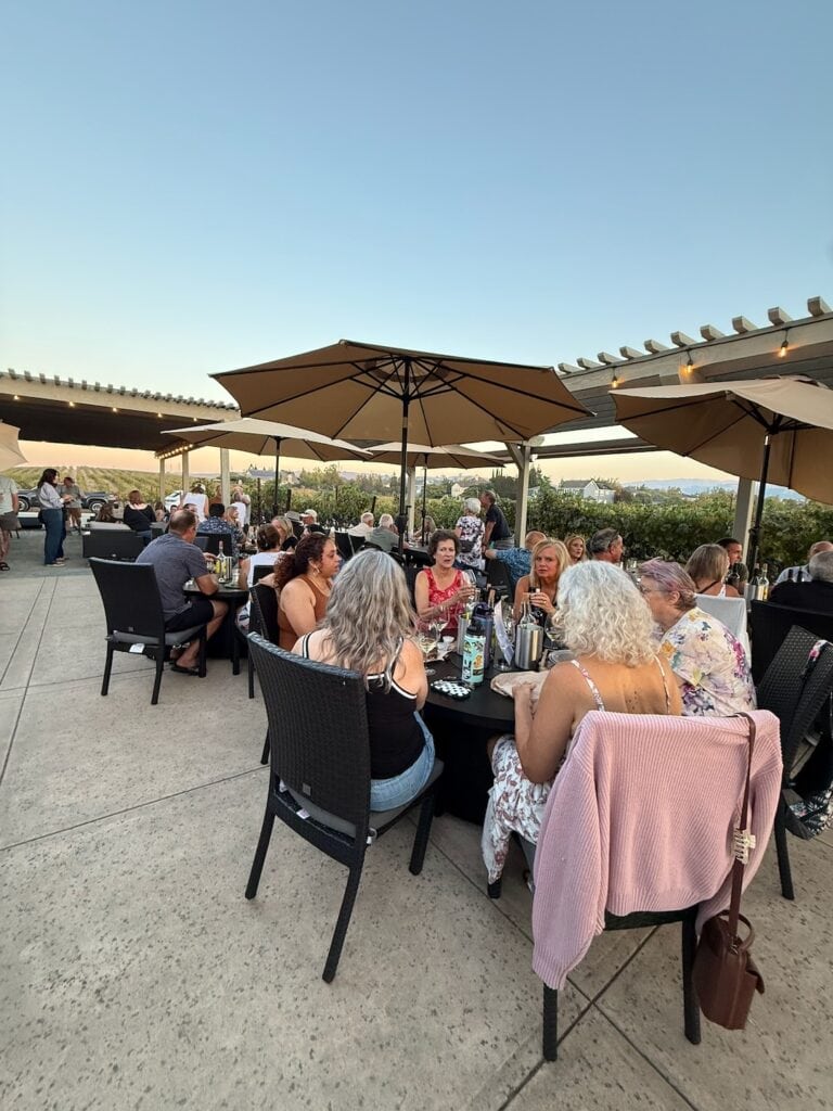 a picture of the Cellar 13 patio filled with people sitting at tables in the evening with the vineyard behind them.